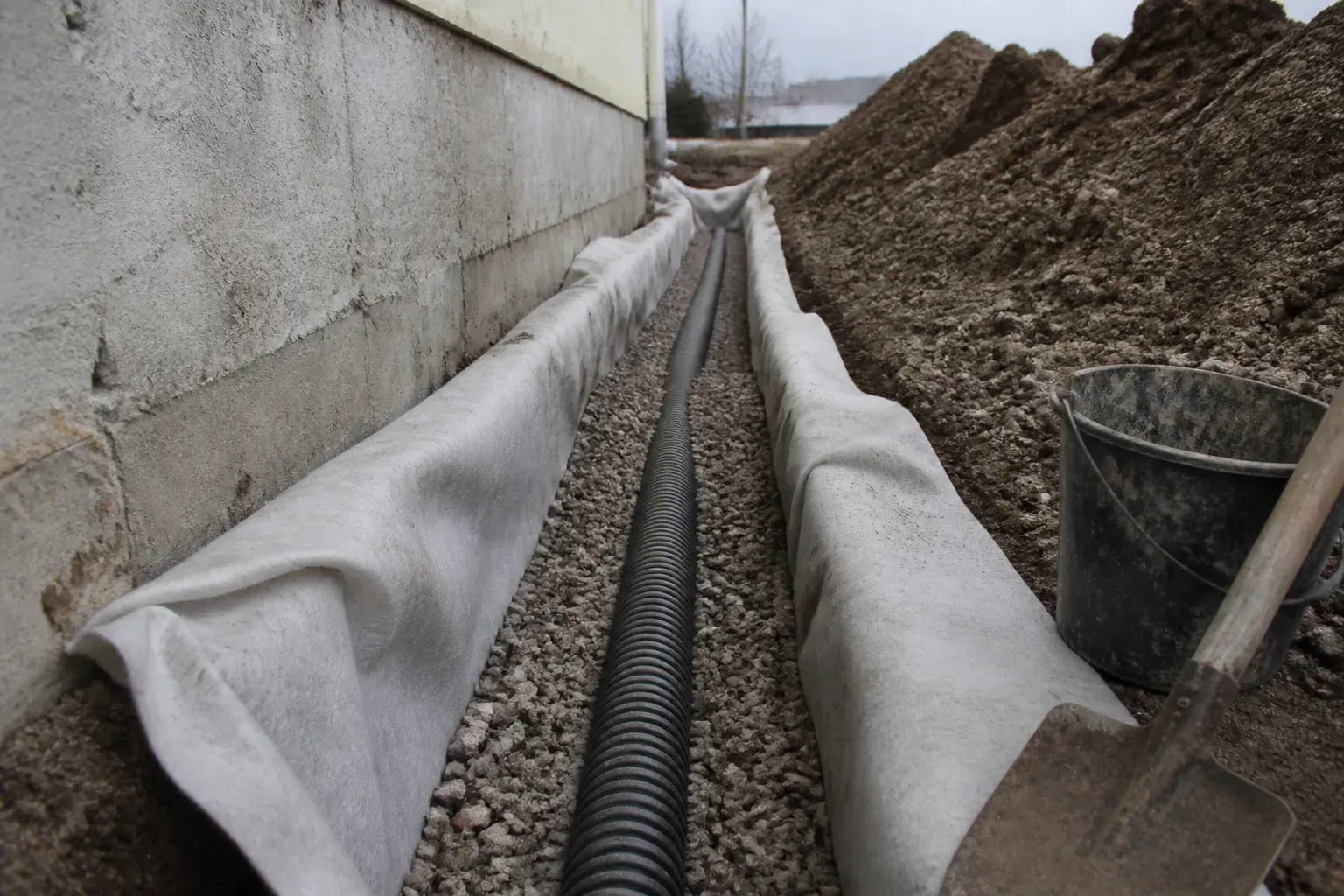 Tranchée de drainage périphérique ouverte le long d’un mur, fond de fouille, drain souple posé et gravier en cours de remblai